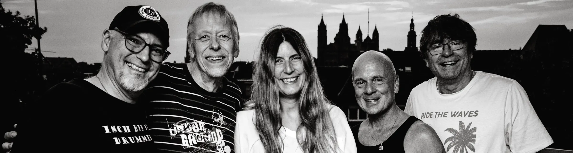 Die fünf Mitglieder von Ready N Rock stehen lachend nebeneinander auf einer Dachterrasse, im Hintergrund die Silhouette des Wormser Doms. Schwarzweiß.