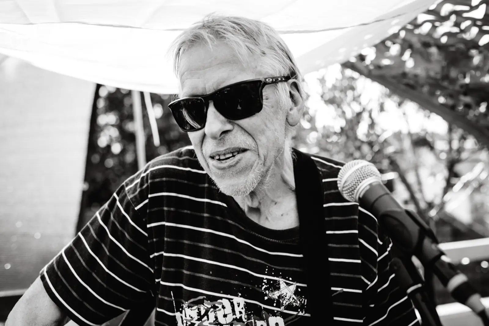 Porträt von Kurt Ludwig mit Sonnenbrille unter dem Sonnensegel, rechts neben ihm das Mikrofon.