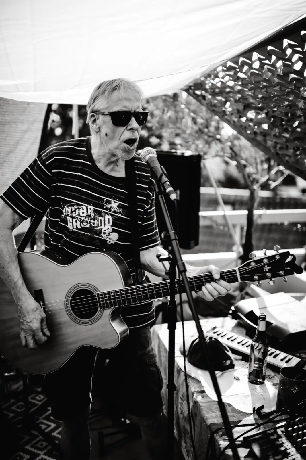 Kurt Ludwig mit Sonnenbrille spielt Akustikgitarre und singt, neben ihm auf dem Tisch ein Keyboard.