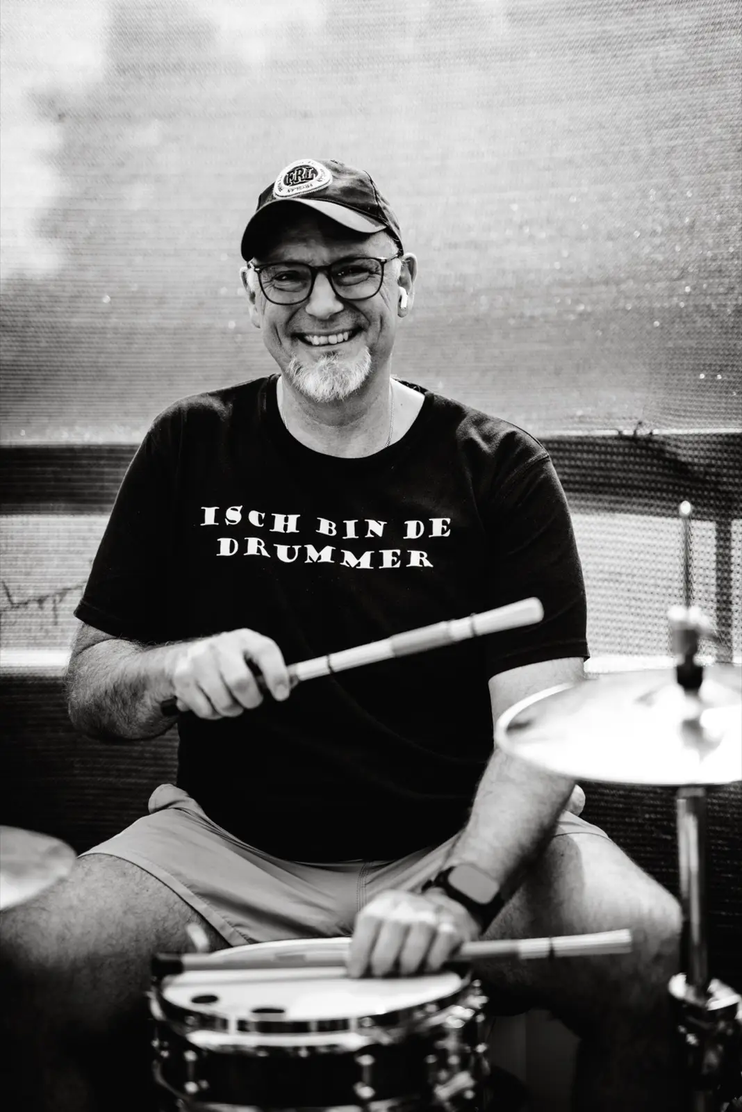 Thomas Südbröcker sitzt lachend am Schlagzeug, mit Cap, Brille und Drumsticks in den Händen; auf seinem T-Shirt steht „Isch bin de Drummer“.